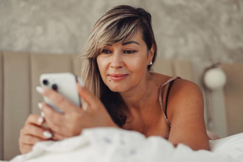 A woman relaxes on her bed while using a smartphone. The soft lighting and cozy bed setting