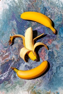 Three bright yellow bananas, one peeled, rest on a vibrant abstract marble background. The colors