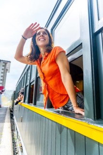 A woman in an orange shirt is waving to someone outside of a train window. Scene is cheerful and