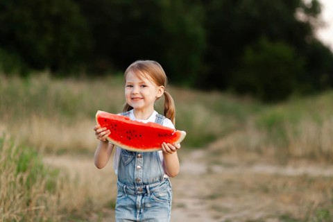 A cheerful child holds a large slice of juicy watermelon, standing outdoors in a lush green field.