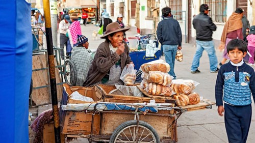 A street vendor with a cart full of bread in a busy street with passers-by and a child, The Uyuni