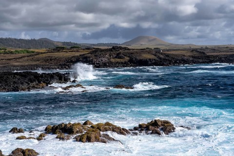 Rocky lava coast with surf and spray, volcanic cones under cloudy sky in the background, Easter
