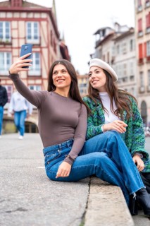 Vertical photo of two stylish friends taking a selfie sitting in a square in the city