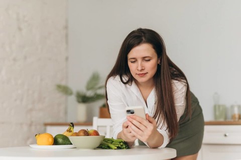 A nutritionist is focused on a smartphone while surrounded by fresh fruits on a kitchen table,