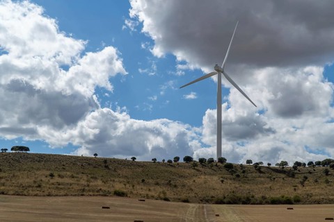 A single wind turbine stands on a dry, hilly landscape under a dynamic sky filled with fluffy