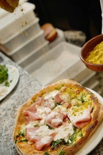 Cropped unrecognizable chef sprinkles seasoning on a freshly prepared pizza topped with ham,