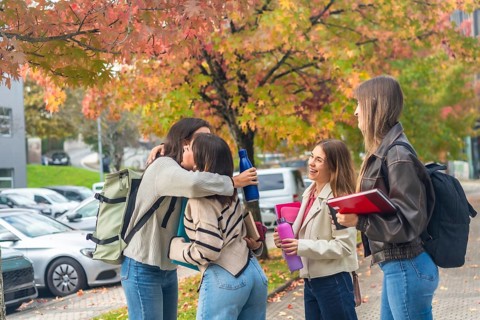 Young female students embracing and laughing on a university campus amid colorful autumn leaves,