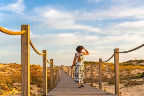 Woman in a long dress walking along a wooden boardwalk path through coastal sand dunes, enjoying a