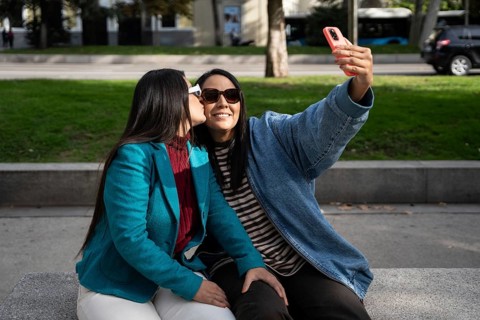 A loving latin lesbian couple sits on a park bench, capturing a joyful selfie. One partner, wearing