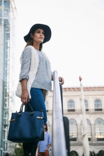 Stylish woman in jeans, sweater, and hat holding a blue handbag, posed thoughtfully in an urban