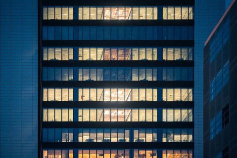 A modern office building illuminated from within as night falls. Rows of bright windows in a city