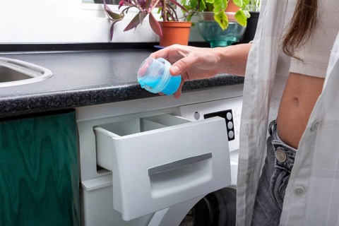 A young woman stands at a front-loading washing machine, holding a blue detergent cap ready to