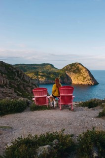 Back view of an unrecognizable woman sitting on pink chairs, overlooking the lush cliffs and