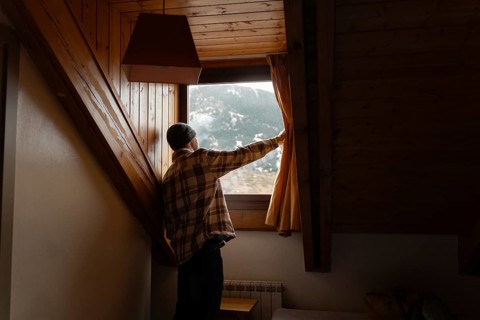 A man in a plaid shirt opens a curtain inside a cozy wooden cabin, revealing a picturesque snowy