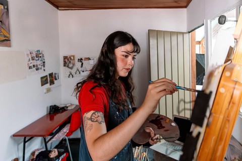 A young woman artist is focused on painting a canvas in her home studio. The cozy space is filled