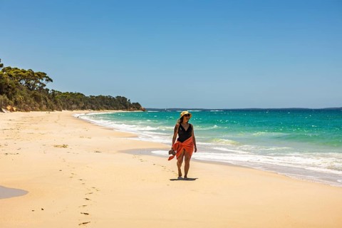 A woman wearing a hat and towel walks along a pristine sandy beach by the turquoise ocean under