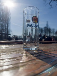 Empty Paulaner beer glass on a weathered beer garden table, behind it beer garden visitors,