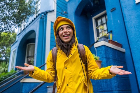 Woman wearing a yellow raincoat and beanie smiling while standing in the rain, hands open to catch