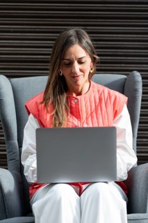 A fashion woman wearing a red and white outfit focuses on her laptop, sitting in a modern armchair.