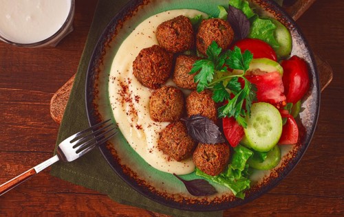 A plate holds falafel balls, tahini, salad with tomatoes and cucumbers, and a drink. The setting is