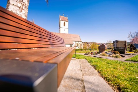 Cemetery with wooden bench and tombstones in front of a church under clear sky, Aidlingen,