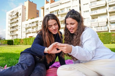 A loving LGBTQ+ young women couple sits on a picnic blanket in a grassy urban park, smiling as they