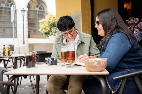 A happy curvy lesbian couple share a joyful moment at an outdoor bar. They write and laugh