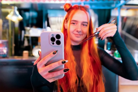A woman with vibrant hair takes a selfie while visiting an Asian food market, holding chopsticks