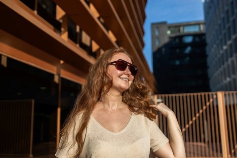 A cheerful woman with long red hair and sunglasses smiles warmly, basking in the sun. Her relaxed