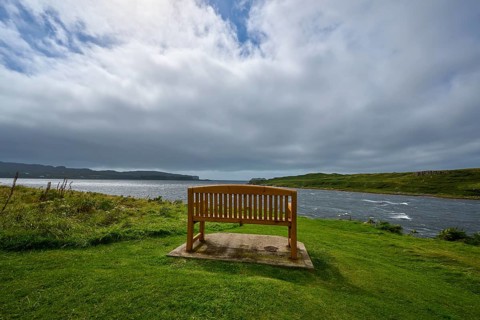 Wooden bench on shore surrounded by green landscape under cloudy sky, Isle of Skye, Scotland,