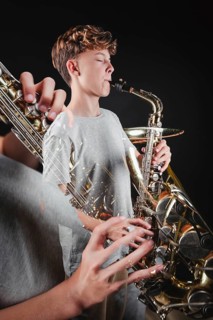 A teenage boy focuses intently as he plays a saxophone in a dark studio setting, highlighting his