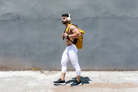 Fit man in white leggings and headphones walking confidently along a textured gray wall, carrying a