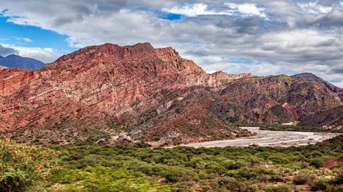 A valley with red rocks and vegetation under a cloudy sky surrounded by mountains, the landscape