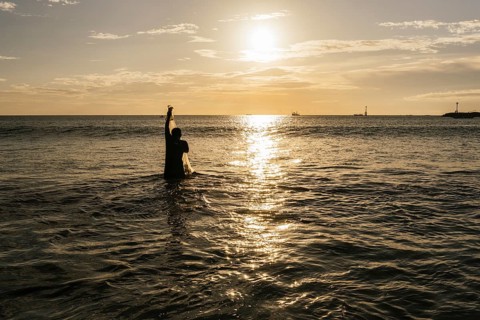 A fisherman stands in the ocean, silhouetted against the golden sunset, casting his net into the