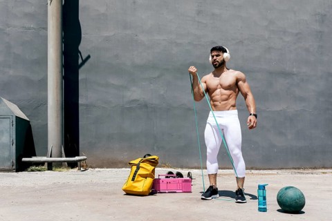 Fit shirtless man with defined muscles performs a bicep curl using a resistance band in an urban