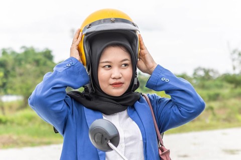 Asian Muslim woman wearing a hijab and a blue jacket, adjusting a yellow helmet, prepares to ride a