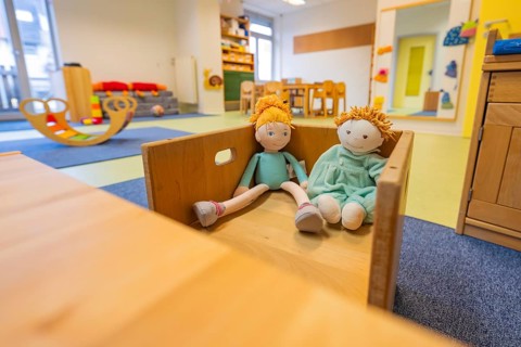 Two dolls sit in a wooden box surrounded by daycare furniture in a room with natural light, infant