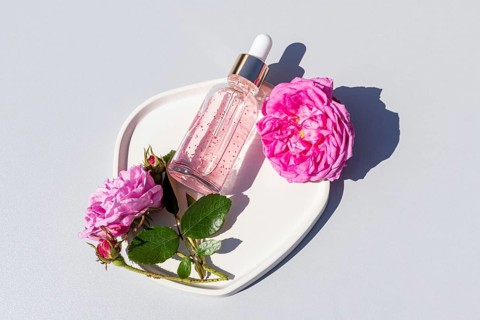 Top view of a clear dropper bottle with rose oil serum, surrounded by vibrant pink roses on a white