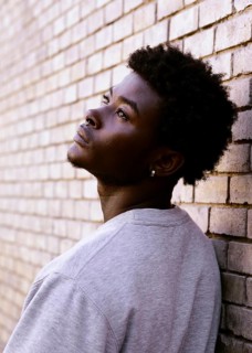 A young black man gazes upward thoughtfully beside a textured brick wall, showing depth and emotion