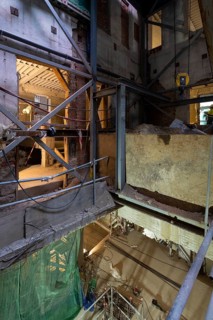 An indoor construction site with metal beams, scaffolding, and workers. The dim lighting highlights