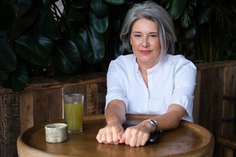 Mature woman sits at a rustic table with a refreshing green drink, surrounded by lush greenery. She