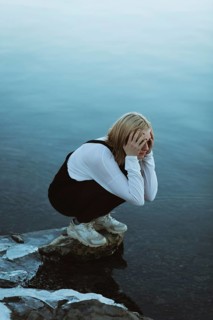 A teenager squats near a calm body of water, with hands covering their face, expressing deep