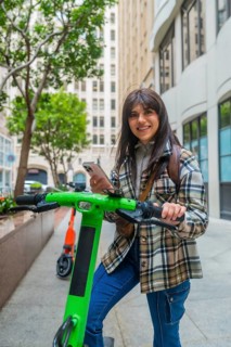Young woman smiling while holding an electric scooter, checking her smartphone to locate or unlock