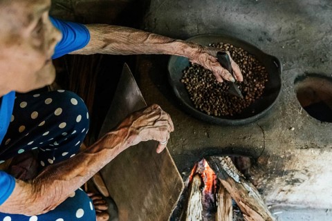An elderly woman roasts coffee beans over an open flame, capturing the essence of traditional