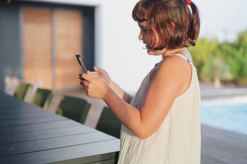 A young girl in a light summer dress stands by the pool, engrossed in her smartphone. The sunny