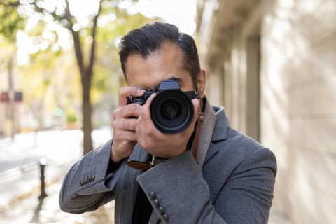 An Indian man, dressed as a photographer with a grey coat, focuses intently on capturing images