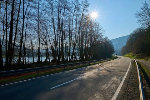 An empty country road lined with trees, with rolling hills in the background and the sun shining