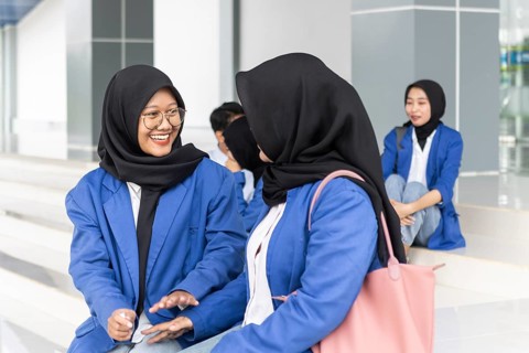 A group of Asian Muslim female students in blue jackets sit together outside a modern school