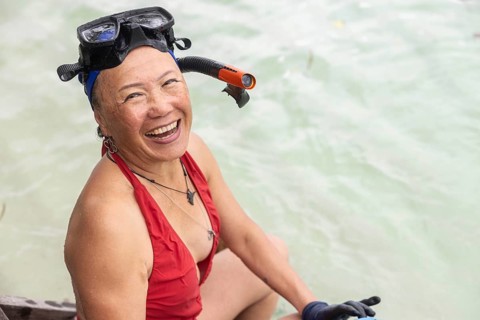 A cheerful elderly Asian woman, dressed in snorkeling gear, sitting by the crystal clear waters of