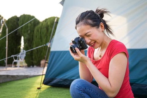 Smiling Asian woman captures moments with her camera in a sunny outdoor setting beside a camping